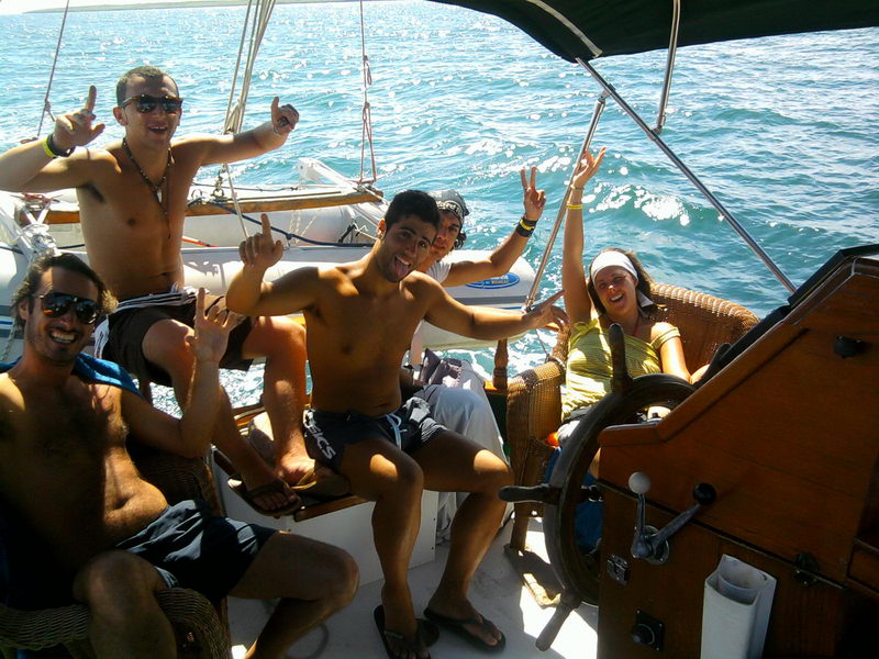 Group of five cheerful young passengers in swimwear posing around the wooden ship's wheel in a sailboat cockpit under a bimini, one woman seated in a wicker chair steering, sparkling sea behind.
