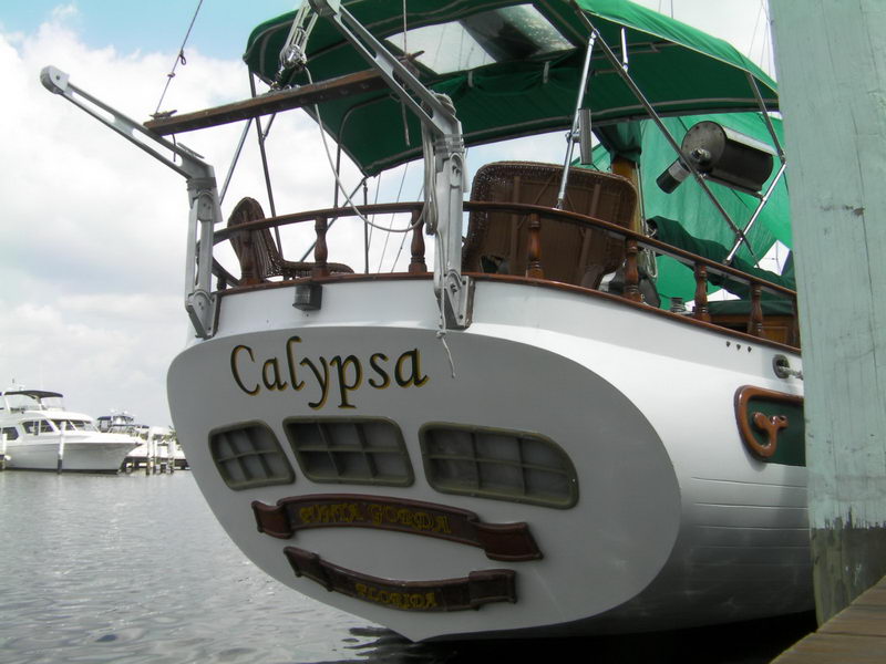Rounded white transom of a classic sailboat named Calypsa from Punta Gorda, Florida, three stern windows, green canvas awning and wicker chairs on the aft deck, boats moored behind on a gray day.