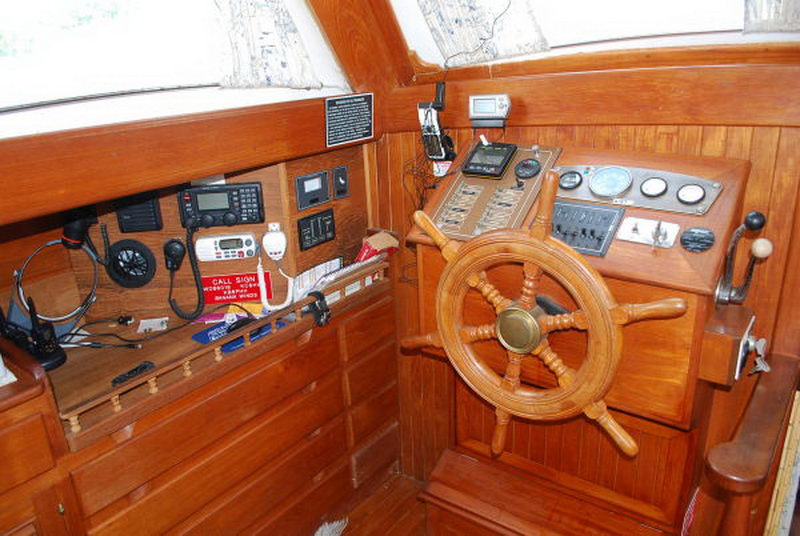 Interior helm station of a classic wooden boat: spoked wooden ship's wheel, varnished mahogany console with engine gauges and switch panel, marine radios, handsets and call-sign card on the left shelf.