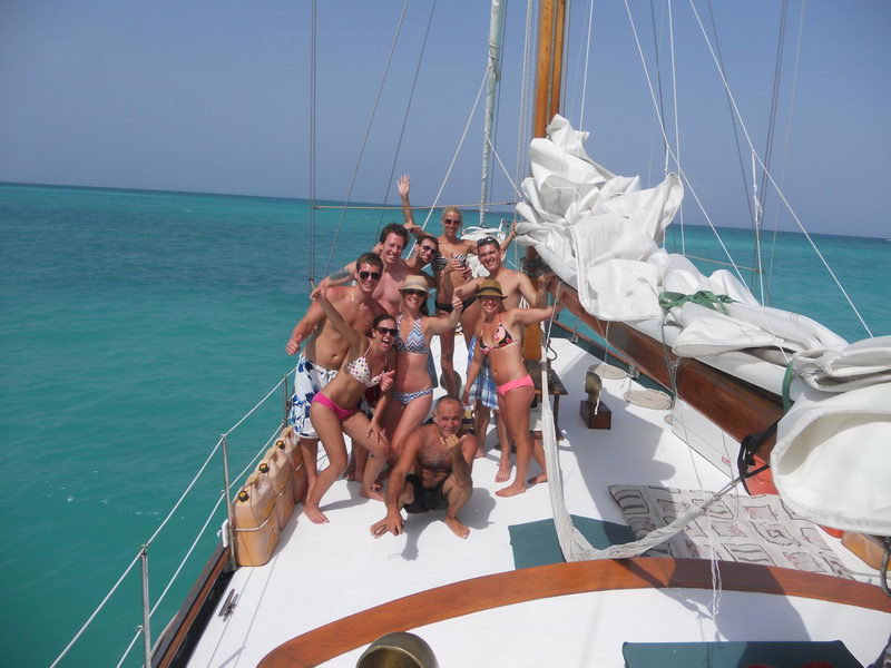 Group of about ten guests in swimwear posing happily on the white foredeck of a classic sailboat with lowered white sails, wooden mast and boom, flat turquoise sea stretching to the horizon.