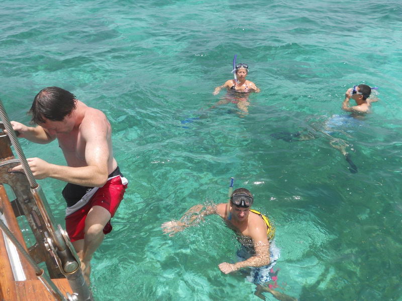 Four snorkelers with masks and snorkels swimming in clear turquoise reef water while a man in red swim trunks climbs down the boat's wooden boarding ladder in the foreground.