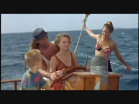 Movie still: a family scene at a sailboat's wooden wheel at sea — a man in a blue cap and a woman steering together, a young boy beside them and a woman in a bikini top holding the rigging.