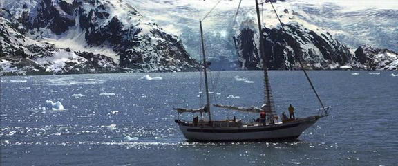 Movie still: a two-masted sailboat motoring through icy water dotted with small ice floes beneath snow-covered mountains and a glacier, crew figures visible on deck.
