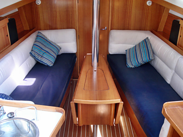 Bright modern sailboat salon with facing white-and-navy upholstered settees, striped accent pillows, drop-leaf wooden table around the mast post, light wood joinery and teak-style flooring.