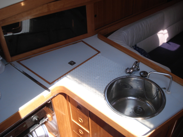 Compact sailboat galley corner with round stainless sink, folding faucet, patterned worktop with cutting-board insert, wooden drawers below and a settee back cushion behind, sunlight streaking in.