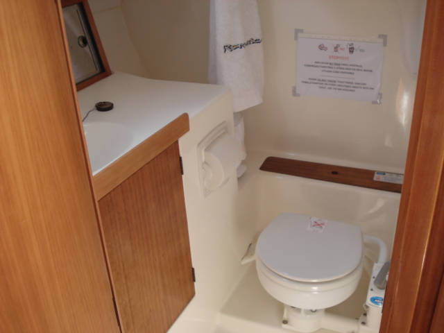 Small white fiberglass sailboat head with manual marine toilet, molded sink counter with wooden cabinet door, mirror, towel hook and an instructions sheet taped to the wall.