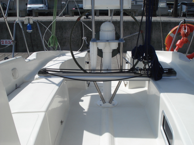 White sailboat cockpit at a dock, large black destroyer-style wheel with steering pedestal and folded cockpit table, white benches, mainsheet traveler across the cockpit sole and an orange life ring aft.