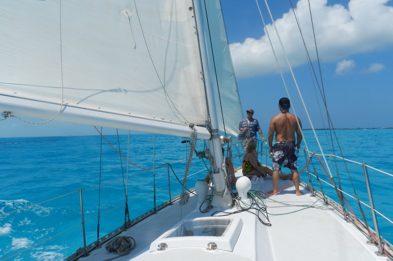Looking forward along a white sailboat's deck under full sail over vivid turquoise sea, four passengers seated and standing near the mast, wake foaming along the port side, puffy clouds ahead.