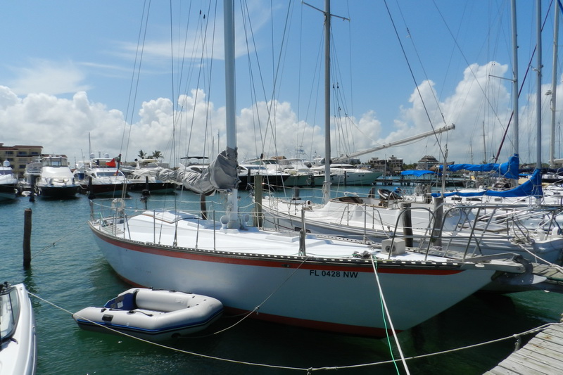 White 44ft monohull sailboat with red waterline stripe docked at a marina, gray inflatable dinghy tied alongside, other yachts and sailboats moored in background under sunny sky with scattered clouds.