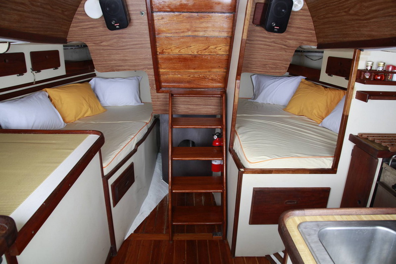 Sailboat interior cabin with two double berths on either side of a wooden companionway ladder, white and yellow pillows, wood paneling, speakers mounted overhead and small galley sink at right.