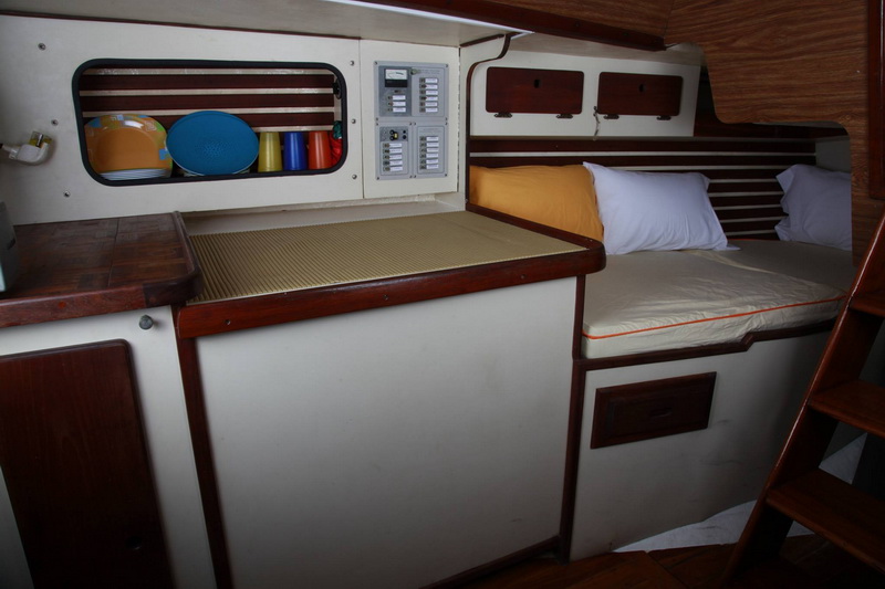 Sailboat interior showing galley counter area with electrical breaker panel, storage cubby with colorful plastic dishware, and a single berth with white and orange pillows against wood-slat trim.