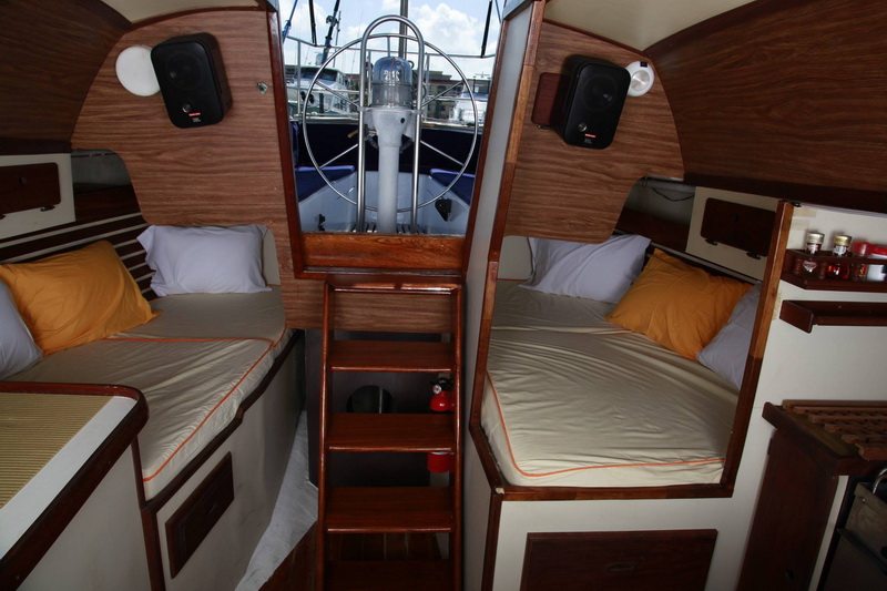 Sailboat interior with two single berths flanking the companionway ladder, yellow and white pillows, wood-grain paneling, and the steering wheel and cockpit visible through the open hatch.