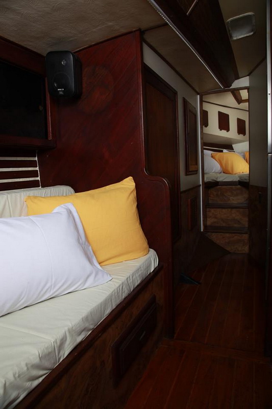 Narrow sailboat interior passageway with settee berth dressed in white and yellow pillows, dark red-brown wood paneling, wall speaker, and another cabin with matching bedding visible through the doorway aft.