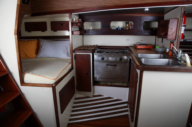 Sailboat galley with stainless steel gimbaled stove/oven, double sink, portable burner, spice rack and storage lockers, with a single berth with orange-trimmed bedding at left and wooden steps in foreground.