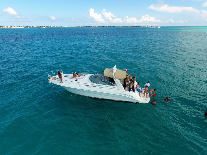 Aerial drone view of a white Sea Ray-style express cruiser anchored in turquoise water with about a dozen guests aboard and several swimming at the stern, tan bimini top, Cancun coastline and reef line in background.