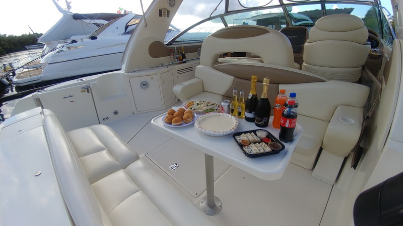 Express cruiser cockpit with cream vinyl seating around a white pedestal table set with sandwiches, sushi trays, champagne bottles and soft drinks, other yachts docked behind under partly cloudy sky.
