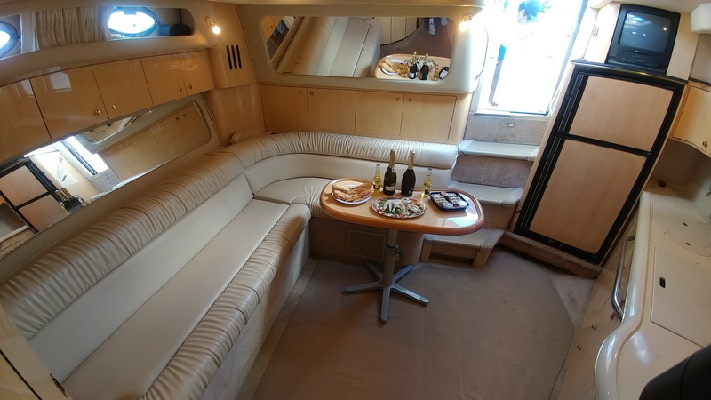 Cabin salon of a cruiser with wraparound pleated cream leather sofa, oval wood table holding champagne bottles, sandwiches and sushi tray, maple cabinetry, mirror panel and small TV, steps up to cockpit.