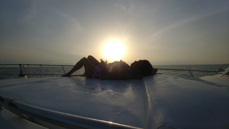 Backlit sunset scene of a woman in a floral swimsuit reclining on a white bow sunpad holding a cocktail glass, sun low over calm sea behind her, bow rails silhouetted.