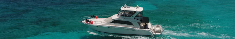 Very wide panoramic banner strip of a white flybridge motor yacht with black hardtop cruising over turquoise reef water, red fenders on bow rail, seen from slightly above.