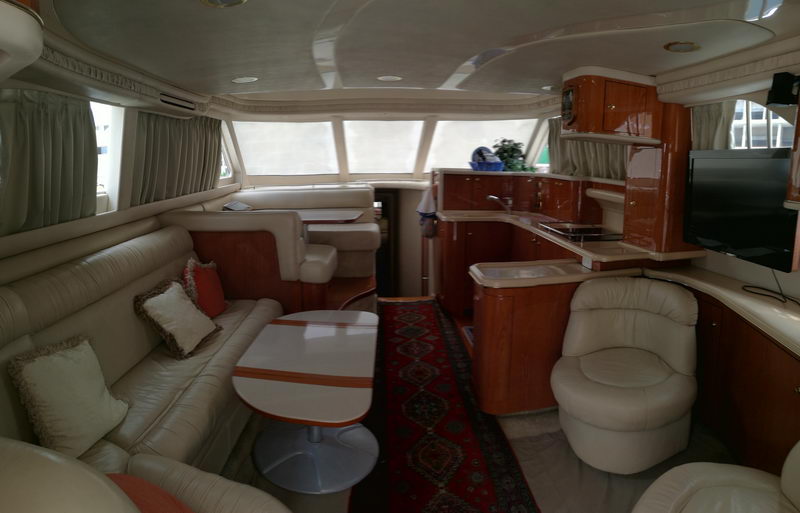 Main salon of a motor yacht with cream leather sofa and armchair, orange-striped oval table, cherry wood galley with sink and cooktop, oriental runner rug, flat TV and curtained windows.