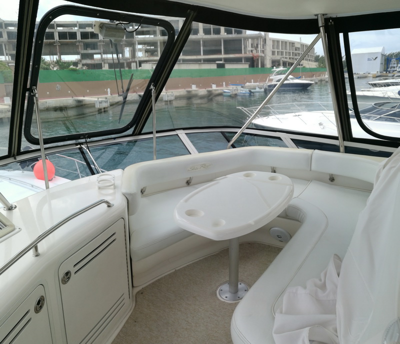 Flybridge of a motor yacht with white vinyl wraparound seating and oval table with cup holders, clear vinyl enclosure panels, marina with docked yachts and an unfinished concrete building behind, overcast light.