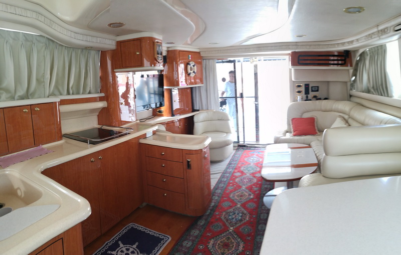 Wide-angle salon view of a motor yacht with cream paneling, cherry cabinetry, corian galley counters with sink, TV on cabinet, patterned oriental rug, and a person entering through the aft glass door.