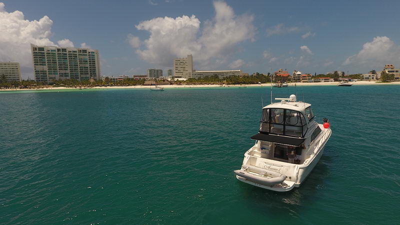 Stern-quarter view of a white flybridge motor yacht anchored in green-blue water off Cancun hotel-zone beach, high-rise hotels and white sand shoreline in background, sunny with scattered clouds.