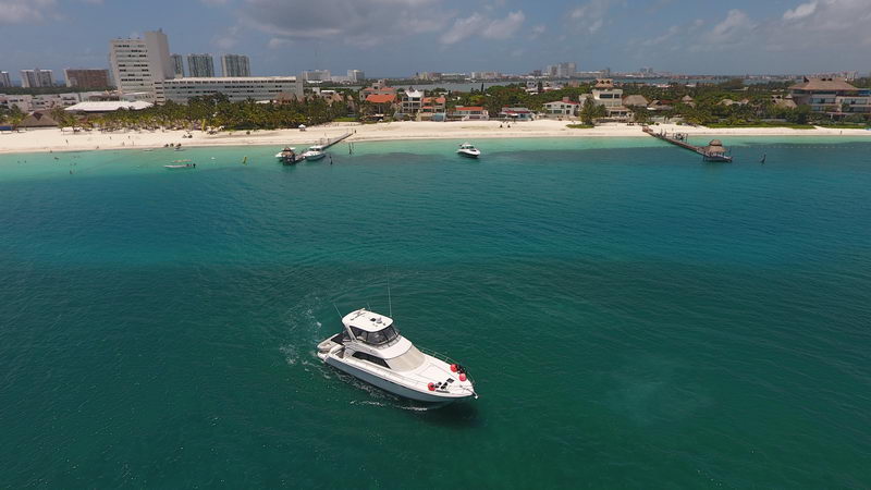 Aerial view of a white flybridge motor yacht anchored in dark seagrass water near a white-sand beach with palapa pier, smaller boats near shore and Cancun hotel skyline on the horizon.