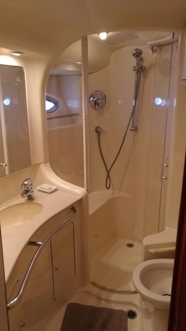 Yacht head interior in cream tones with molded vanity sink, mirror, oval porthole window, separate shower stall with handheld chrome fixture and glass door, toilet at right.