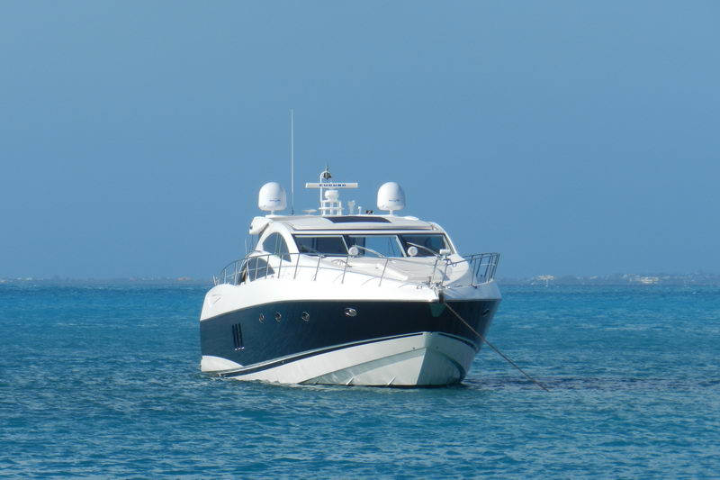 Bow-quarter view of a navy-hulled Sunseeker sport yacht at anchor with mooring line running from the bow, twin satellite domes and radar mast above the enclosed hardtop, hazy horizon over blue water.