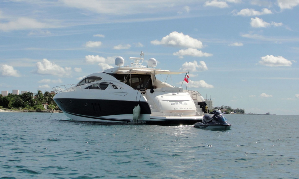 Stern-quarter view of dark-hulled sport yacht Aguila anchored under a partly cloudy sky, canvas awning over the cockpit, Mexican flag at stern, black and blue jet ski tied alongside the swim platform, green coastline behind.