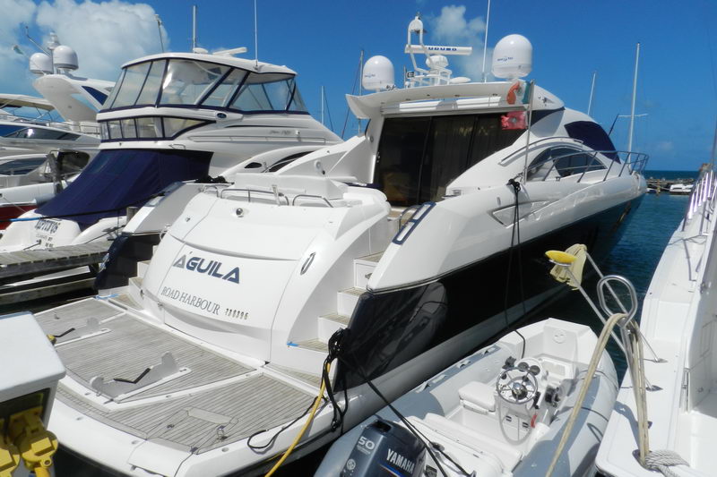 Stern of sport yacht Aguila (Road Harbour) berthed in a marina, teak swim platform and boarding steps, mooring lines and yellow bollard, neighboring motor yachts and outboard tender around, sunny blue sky.