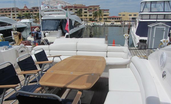 Aft cockpit of a sport yacht with oval teak table, white L-shaped sofa and navy folding director chairs, teak deck, marina behind with docked yachts and Mediterranean-style waterfront buildings.