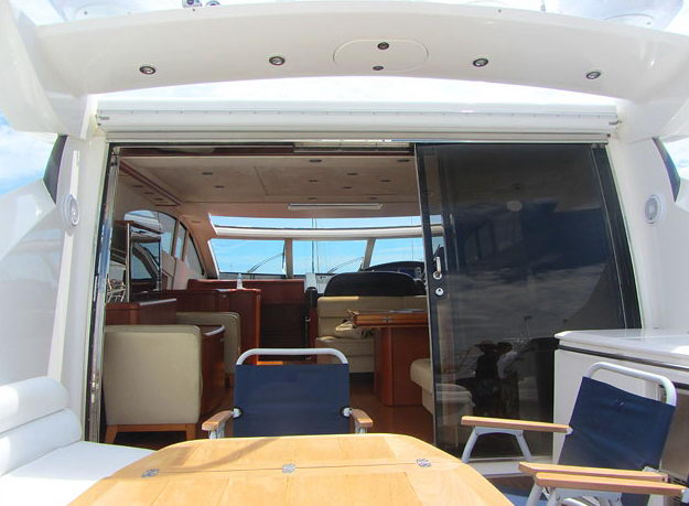 View from the shaded aft deck through a wide sliding glass door into the salon of a sport yacht, cream leather armchairs, cherry wood tables and helm area beyond, navy deck chairs and teak table in foreground.