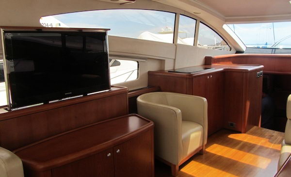 Salon of a sport yacht with pop-up TV raised from a cherry wood sideboard, cream leather tub chair, wood flooring and cabinetry, curved windows overlooking a marina with boats and masts.