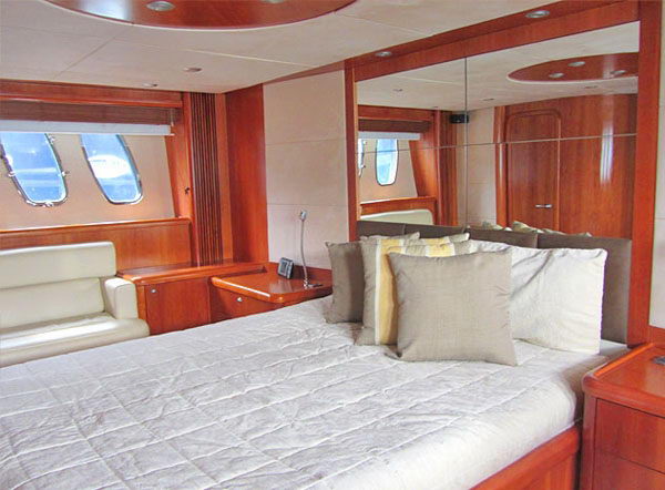 Interior yacht master cabin with glossy wood paneling, mirrored wall, two portholes, an off-white leather bench and a made-up bed with cream and taupe pillows.