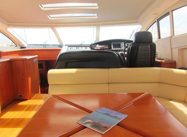 Interior helm and salon area of a motor yacht, showing the black leather helm seat and dashboard, cream leather bench seating, and a wooden dining table with a magazine on top.