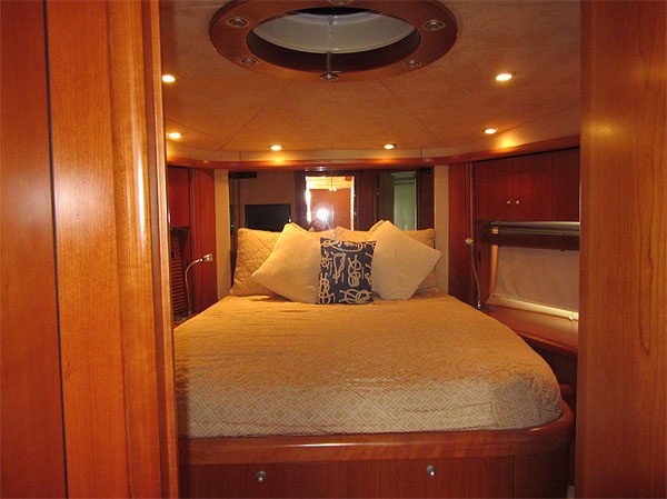 Interior yacht cabin bed viewed through a doorway, with wood paneling, recessed ceiling lights, cream bedspread and a navy patterned throw pillow among white pillows.