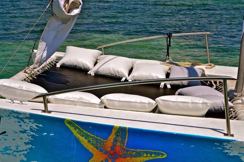 Close-up of a catamaran's trampoline deck with white cushions, a furled sail, and a painted orange-and-yellow starfish design on the blue hull, green water below.