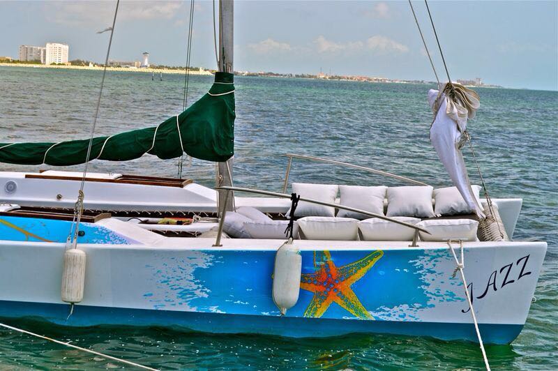 Close-up of the 'Jazz' catamaran's bow at anchor with a green sail cover, painted starfish hull design, and the Cancun skyline visible across the water.