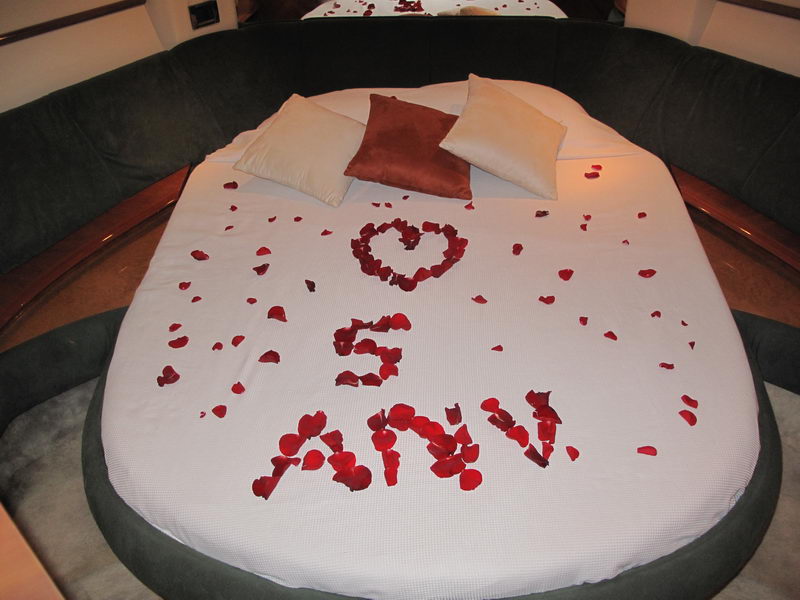 Yacht interior cabin showing a round berth decorated with red rose petals arranged in a heart shape and the text '5 ANIV', cream and dark brown accent pillows against dark upholstered headboard, romantic anniversary setup.
