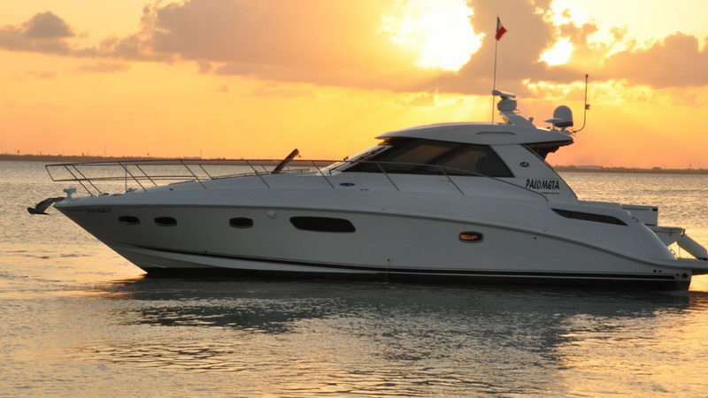 Sport motor yacht at rest on calm water during sunset, side view with a Mexican flag flying from the stern, orange sky and its reflection covering the water surface, hull name visible.