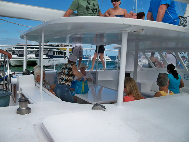 Catamaran deck crowded with passengers docked at a marina, other tour boats moored nearby, people boarding and socializing under a partly cloudy sky.