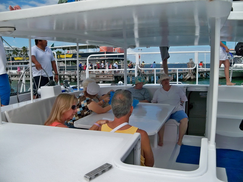 Catamaran deck with a group of tourists, including older adults, seated together while the boat is moored near a dock; other catamarans and a canopy structure visible in the background under an overcast sky.