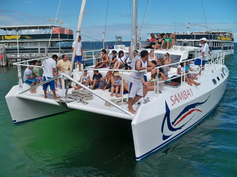 Catamaran named 'Samba 1' sailing with its mainsail raised, deck full of passengers sunbathing and seated, moored close to other tour boats near a marina under a cloudy sky.