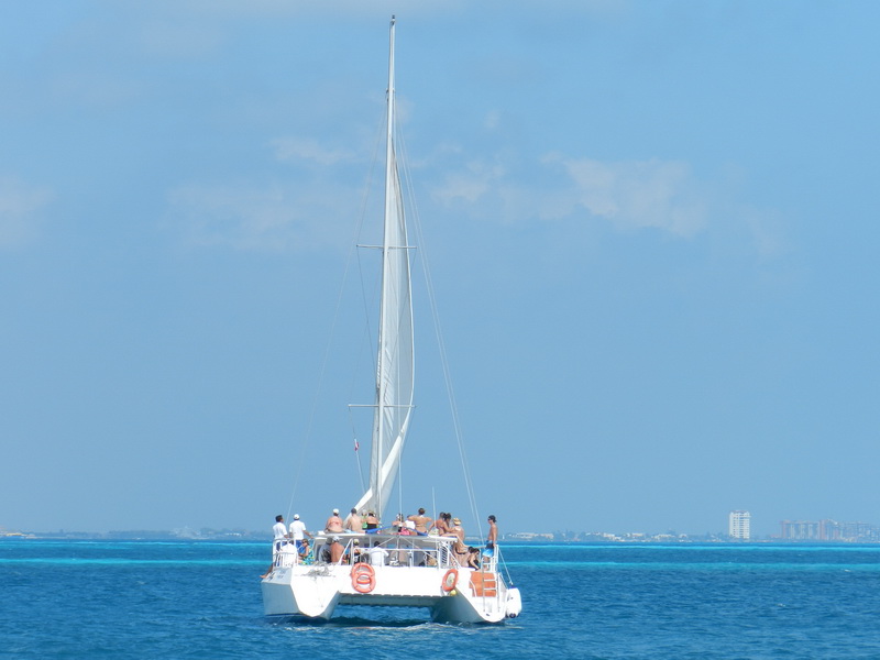 Catamaran named 'Samba' sailing under full sail on open turquoise sea with passengers visible on deck, a distant city skyline on the horizon under a sunny sky.
